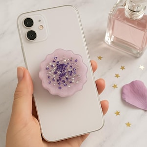 May include: A light purple phone grip with a floral design, adorned with silver and purple crystals, is attached to a white smartphone. The phone is held in a hand, with a perfume bottle and decorative stars in the background.