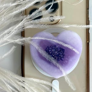 May include: A heart-shaped phone grip in shades of purple, adorned with a cluster of dark purple crystals at its center. The grip is mounted on a clear phone case, with white feathers in the foreground.