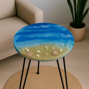 Pode incluir: Mesa lateral redonda com tampo em resina epóxi com tema de praia. O tampo apresenta um design de céu azul e branco, uma praia arenosa e conchas embutidas. A mesa tem pernas de hairpin pretas e está sobre um tapete de juta tecido.