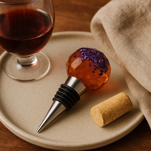 Puede incluir: Un tapón decorativo para botella de vino con base de acero inoxidable y una parte superior de globo transparente de color naranja con purpurina morada. Una copa de vino y un corcho también están en el plato.