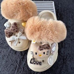 May include: Beige clogs with brown fur trim. One has a bow and heart, the other a cartoon cat and the word "Love". The clogs are decorated with various charms and embellishments.