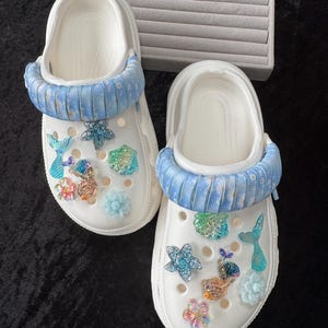 May include: White clogs with blue fabric straps and decorative charms. The clogs are decorated with mermaid tails, starfish, and floral embellishments. These casual shoes are ideal for a beach or summer theme.