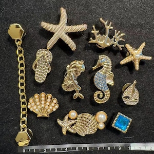 May include: A collection of gold-toned nautical-themed metal pins and charms, including a starfish, seahorse, seashell, sailboat, and flip-flop, some accented with faux pearls, blue crystals, and a chain, measured by a ruler in centimetres.