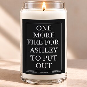 May include: A clear glass jar candle with a white wax fill and a lit wick. The black rectangular label features white text that reads: ONE MORE FIRE FOR ASHLEY TO PUT OUT. The bottom of the label includes the text: Burn: 60-80 HRS, Surprised Me, 100% SOY WAX.