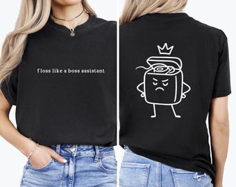 May include: Black t-shirt with the text "floss like a boss assistant" in white. The back of the shirt features a white cartoon illustration of a floss container with a crown, arms, and legs, displaying an angry expression.