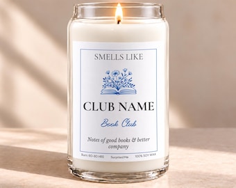 May include: A lit candle in a clear glass jar with a white label featuring blue text and a floral book illustration. The label reads: SMELLS LIKE CLUB NAME Book Club Notes of good books & better company. Burn: 60-80 HRS 100% SOY WAX.