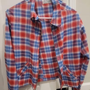 May include: A red, white, and blue plaid jacket with a zipper closure and two front pockets. The jacket is hanging on a white hanger against a white door. The jacket has a collar and long sleeves.