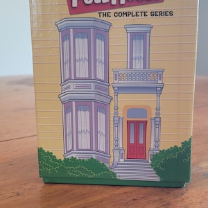 Può includere: Un cofanetto giallo di "Full House: The Complete Series" con un'illustrazione a cartoni animati dell'iconica casa. La scatola presenta il logo dello show in rosso e bianco e l'illustrazione della casa ha accenti viola e rossi.