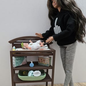 May include: A doll stands next to a brown changing table with a baby doll lying on top. The changing table has shelves with baby products, including diapers and a blue soap dispenser. The doll is wearing a black hoodie and gray pants.