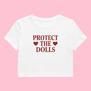 May include: White crop top featuring the maroon text "PROTECT THE DOLLS" with two heart symbols. The top has a crew neck and short sleeves. The shirt is made of a soft material.