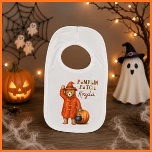 May include: A white baby bib featuring a graphic of a brown bear wearing an orange coat and a matching pointed hat. The bear stands next to a pumpkin and holds a small black cauldron. The text reads Pumpkin Patch and the name Kayla in brown.