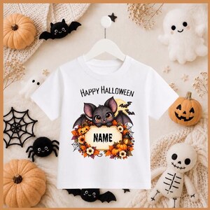 May include: A white short-sleeve t-shirt featuring a graphic of a cute bat with large eyes, pumpkins, autumn leaves, and a lantern. The text "Happy Halloween" is printed above the bat, and the word "NAME" is centred in a decorative frame.