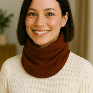 May include: A woman wearing a dark red neck warmer. The neck warmer is made of a soft, knitted material. She is wearing a cream-colored ribbed sweater. The background is a neutral color.