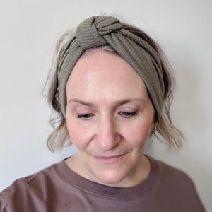 Peut inclure: Une femme porte un bandeau en tricot c&ocirc;tel&eacute; vert sauge avec un n&oelig;ud sur le dessus. Le bandeau est fait d'une mati&egrave;re douce et extensible et est con&ccedil;u pour maintenir les cheveux hors du visage. La femme porte un haut marron.