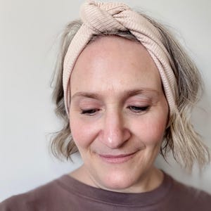 Peut inclure: Un bandeau en tricot c&ocirc;tel&eacute; beige clair avec un dessus nou&eacute;. Le bandeau est port&eacute; sur la t&ecirc;te. La personne sur l'image porte un haut marron.