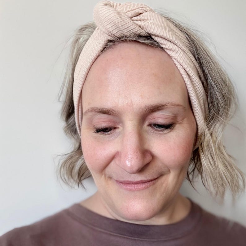 Peut inclure: Un bandeau en tricot c&ocirc;tel&eacute; beige clair avec un dessus nou&eacute;. Le bandeau est port&eacute; sur la t&ecirc;te. La personne sur l'image porte un haut marron.
