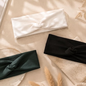 May include: A set of three fabric headbands featuring a twisted knot detail at the centre. The headbands are available in solid white, solid black, and solid dark green colours. Each headband has a wide, rectangular shape with soft edges.