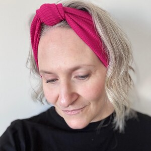 Peut inclure: Un bandeau en tricot c&ocirc;tel&eacute; magenta avec un design nou&eacute;. Le bandeau est port&eacute; sur la t&ecirc;te. La personne porte un haut noir. L'arri&egrave;re-plan est un mur blanc uni.