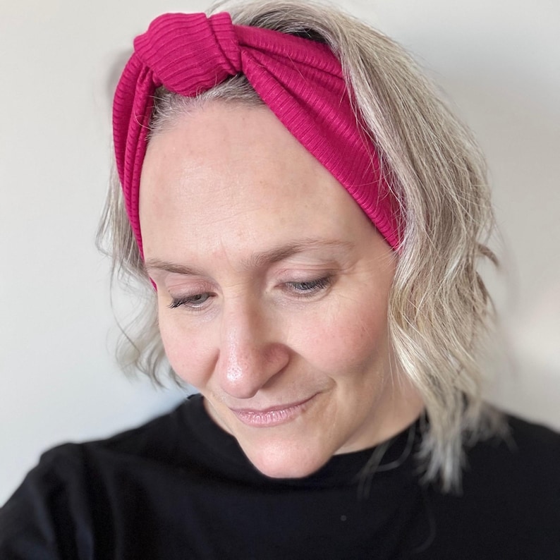 Peut inclure: Un bandeau en tricot c&ocirc;tel&eacute; magenta avec un design nou&eacute;. Le bandeau est port&eacute; sur la t&ecirc;te. La personne porte un haut noir. L'arri&egrave;re-plan est un mur blanc uni.