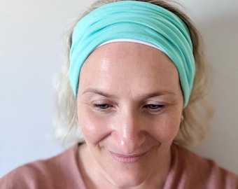 May include: A wide, solid turquoise headband featuring a soft, gathered fabric construction. The headband is positioned across the forehead, displaying a smooth, matte texture and a consistent, vibrant color throughout the entire accessory.