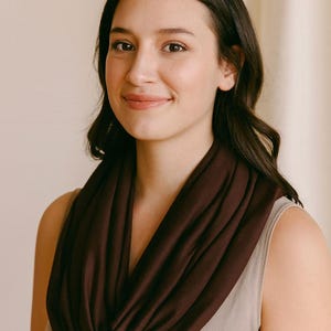 May include: A dark burgundy infinity scarf draped around the neck. The scarf is made of a soft, flowing fabric. The model is wearing a light beige tank top. The background is a neutral color.