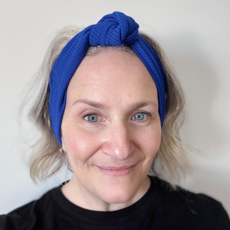 Peut inclure: Un bandeau en tricot c&ocirc;tel&eacute; bleu roi nou&eacute; sur le dessus. Le bandeau est port&eacute; sur la t&ecirc;te. La personne sur l'image porte un haut noir.
