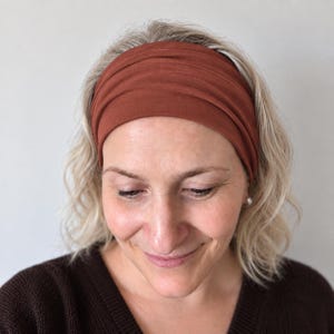 May include: A wide, solid rust-coloured fabric headband with a smooth, matte texture. The headband features a simple, seamless design that wraps around the head, providing a clean and uniform appearance across the front and sides of the accessory.