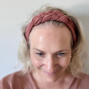 May include: A rose-coloured braided headband. The headband has a thick, woven design across the top and a smooth band. The fabric looks soft and comfortable, suitable for daily wear. The colour is a muted, earthy tone.