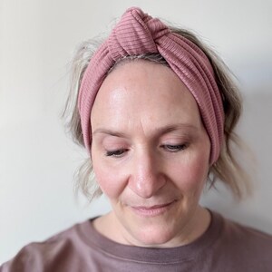 Peut inclure: Un bandeau en tricot c&ocirc;tel&eacute; rose poudr&eacute; avec un n&oelig;ud sur le dessus. Le bandeau est large et couvre le front. Le tissu est c&ocirc;tel&eacute; verticalement. La personne qui porte le bandeau regarde vers le bas.