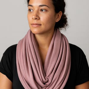 May include: A dusty rose infinity scarf made of soft fabric. The scarf is draped around the neck, creating multiple layers. The person in the image is wearing a black t-shirt. The background is a solid light gray.