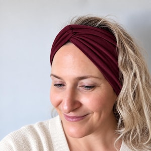 May include: A deep burgundy headband featuring a twisted knot detail at the centre front. The fabric has a soft, matte texture and is worn across the forehead, showcasing a wide, gathered design that wraps around the head with a solid colour.