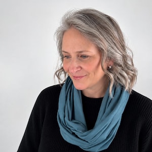May include: A solid blue infinity scarf draped in multiple loops around the neck, worn over a black long-sleeved jumper. The scarf features a smooth, matte fabric texture with soft folds and a consistent deep blue colour throughout the material.