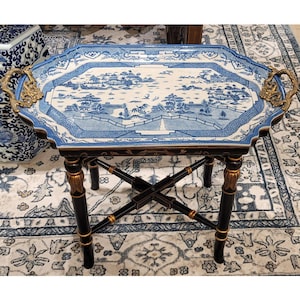 May include: An octagonal accent table with a blue and white landscape design on the tabletop. The table has black legs with gold accents and ornate brass handles. The table is placed on a patterned rug.