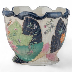 May include: A decorative ceramic planter with a scalloped rim and a flared base. The planter features a cream-colored background with large leaf designs in shades of green, pink, and navy blue, accented with gold trim and floral details.