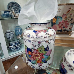 May include: A cylindrical ceramic tissue holder with a white base featuring a colorful floral pattern of pink, orange, and green leaves. The top opening is rimmed in dark blue, and a white tissue is protruding from the center of the container.