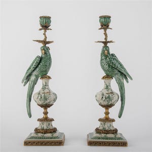 May include: Pair of decorative candle holders featuring green parrot sculptures atop ornate, white and gold bases. The candle holders have a square base and are accented with gold and green details.