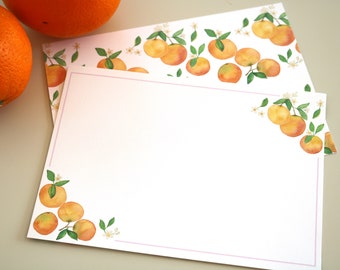 May include: A blank white card with a pink border and a watercolour illustration of orange blossoms and oranges in the corners. The card is on a light brown surface.