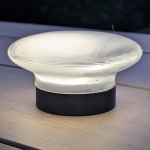 Pode incluir: Uma pequena lâmpada de exterior iluminada com um topo branco em forma de cogumelo e uma base preta. A lâmpada está sobre uma superfície de madeira, com um fundo desfocado de uma cerca e folhagem. A lâmpada emite um brilho suave e quente.