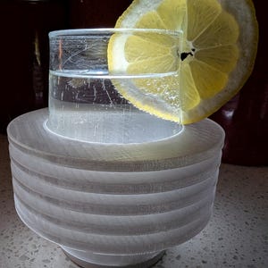 May include: A clear glass filled with water and a lemon slice, resting on a white, layered, circular stand. The stand has a series of horizontal ridges. The drink is set against a dark background.