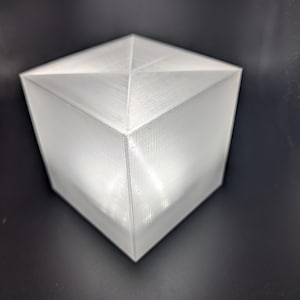 May include: A white, geometric cube with a pyramidal top. The cube has a textured surface and is set against a dark background. The design is modern and minimalist.