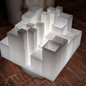 Pode incluir: Uma escultura branca, geométrica e iluminada que se assemelha a uma paisagem urbana. O design apresenta formas retangulares e quadradas em camadas, criando uma estética arquitetônica moderna. A fonte de luz é interna.