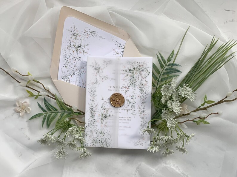 May include: A white wedding invitation with a green floral design and a wax seal. The invitation is on a white background with a white sheer fabric overlay.