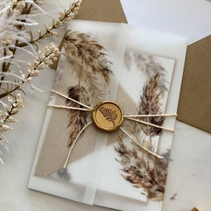 Puede incluir: Un conjunto de invitaciones de boda con una tarjeta color crema con un diseño de hierba de la pampa en acuarela. La invitación está envuelta en vitela y atada con hilo, sellada con un sello de cera dorado. Se ve un sobre marrón en el fondo.