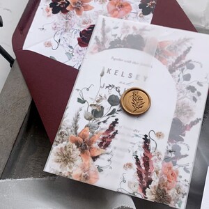 Puede incluir: Una invitación de boda blanca con un diseño floral en tonos rosa, rojo y marrón. La invitación presenta un sello de cera dorado con un diseño de hoja y el texto "Kelsey and".