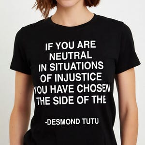 Puede incluir: Camiseta negra con texto blanco que cita a Desmond Tutu: "SI ERES NEUTRAL EN SITUACIONES DE INJUSTICIA, HAS ELEGIDO EL LADO DE..." La camiseta es de cuello redondo y manga corta.