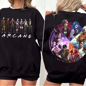 May include: Black sweatshirt featuring characters from the animated series "Arcane" on the front and back. The front displays the word "Arcane" in a stylized font with character illustrations above. The back showcases a circular collage of additional characters.
