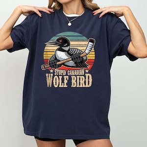 May include: Navy blue t-shirt featuring a cartoon loon bird holding a hockey stick against a sunset background. The text "STUPID CANADIAN WOLF BIRD" is printed below the graphic in a distressed font. The shirt has a crew neck.