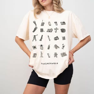 Könnte beinhalten: Cremefarbenes T-Shirt mit einer Anordnung von schwarz-weißen Illustrationen von Geisterjäger-Ausrüstung. Das Wort "PHASMOPHOBIA" ist unten in Schwarz aufgedruckt. Das T-Shirt wird von einer Person getragen.