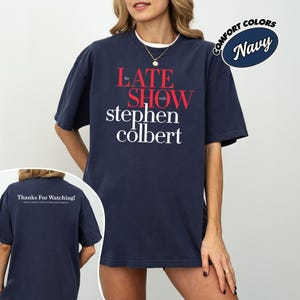 May include: Navy blue t-shirt with the text "The Late Show with Stephen Colbert" in red and white. The shirt has a crew neck and short sleeves. The words "Comfort Colors" and "Navy" are in a circular design.