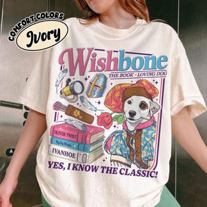 May include: An ivory t-shirt featuring a graphic of a dog dressed in a Renaissance-style hat and tunic holding a rose. The design includes a telescope, a quiver of arrows, and a stack of books labeled Oliver Twist, Rip Van Winkle, and Ivanhoe.